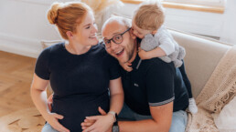 Lächelnde Familie auf einer Couch: schwangere Frau, Mann und Kind, die sich mit einem Stofftier umarmen. -fotostudiorerich