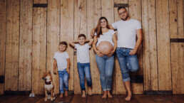 Eine barfüßige Familie mit einer schwangeren Frau und einem Hund posiert lächelnd an einer Holzwand. -fotostudiorerich